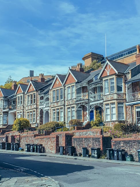 A row of Victorian-style terraced houses with ornate white ironwork balconies and large bay windows. The buildings feature brick facades with decorative trim, gabled roofs, and small front gardens enclosed by low brick walls. In front of the houses, there is a paved sidewalk and a street with black wheelie bins lined along the curb. The sky above is clear with a few wispy clouds, and some greenery from trees and shrubs is visible in the gardens and surrounding areas, set against a backdrop of taller modern buildings. The scene depicts a residential neighbourhood suitable for private waste collection or local rubbish disposal services, with no visible litter or waste but a typical setup of refuse bins for waste management.