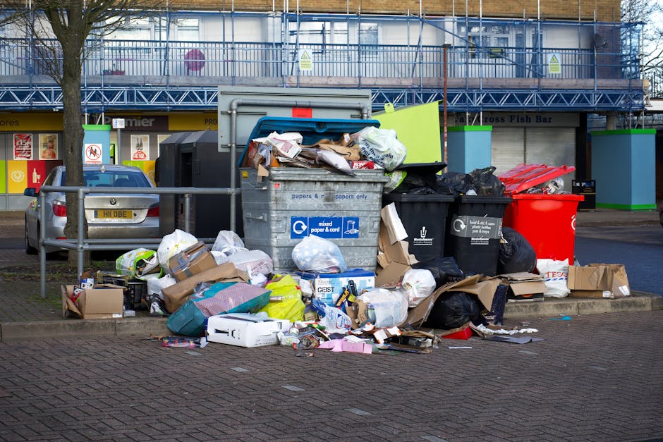 A large collection of mixed waste materials is piled on a paved area in front of a row of waste containers, with some bags and cardboard boxes spilling onto the street. The waste includes various black garbage bags, crumpled cardboard boxes, plastic packaging, and paper waste. The containers are positioned behind the pile, with a central grey recycling bin labeled for mixed paper and card, partially overflowing with paper and packaging. To the right, black and red waste bins are visible; the black one appears to contain general waste, while the red one may be designated for specific disposal. On the left, a parked silver car with a UK license plate is situated next to a metal railing and a small tree, with a background that features storefronts with bright signage and a building with scaffolding and blue safety netting, indicating ongoing construction or renovation. The scene suggests an areas of informal overflow from local waste disposal, highlighting the need for efficient rubbish removal services or private collection options in the vicinity.