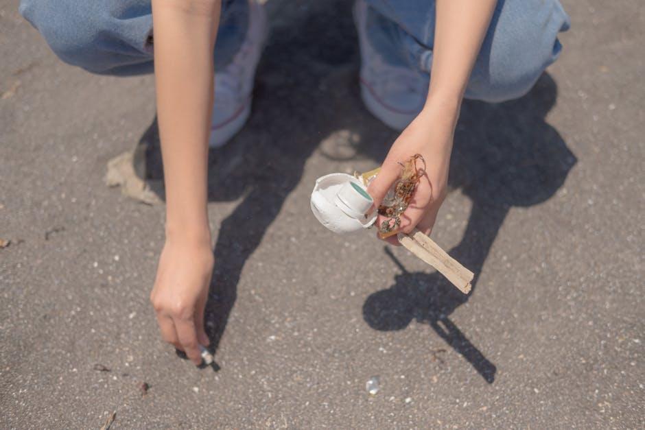 A close-up of a person's hands and knees as they crouch on a paved outdoor surface, holding a cluster of discarded items including a partially broken white plastic cup, tangled pieces of string or rope, and small fragments of debris. The person is wearing light-colored jeans and white sneakers with red accents. The background shows the textured gray asphalt ground with a few small pieces of litter scattered around. This scene illustrates the importance of rubbish removal and the necessity for proper disposal of waste materials, often handled through private collection services. The natural lighting highlights the textures of the debris and the pavement, emphasizing the need for efficient debris clearance to maintain cleanliness and environmental standards.