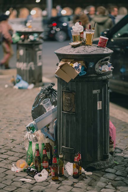 A black, cylindrical rubbish bin made of metal with vertical ridges stands on a cobblestone street in an urban environment, surrounded by scattered litter including broken glass bottles, crumpled paper, and food containers at its base. On top of the bin, there are several used paper cups with plastic lids, some containing remnants of beverages, along with a discarded snack wrapper and a small cardboard box. The background features a blurred view of other cars parked along the street, as well as a grey dustbin and a collection of unidentified objects, indicating an area where waste accumulates and requires collection or clearance. The scene captures typical urban littering, emphasizing the importance of rubbish removal services for maintaining cleanliness in public spaces.