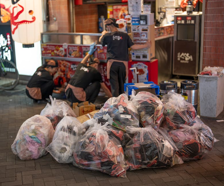 An outdoor scene showing several large, clear plastic garbage bags filled with mixed waste, including clothing, textiles, and miscellaneous items, stacked on a brick-paved surface. Behind the bags, three workers dressed in black uniforms with brown aprons and gloves are actively engaged in sorting or loading additional rubbish. They are situated near a shop or food establishment with red and white signage, illuminated by ambient lighting, indicating it is likely evening or nighttime. The environment includes various waste disposal containers, such as metal bins and a grey plastic trash can, positioned against the building wall. In the background, there are stacked cardboard boxes and small appliances, suggesting a collection or disposal process related to local rubbish removal services. This scene illustrates a typical setup for private rubbish collection or on-site clearance, highlighting the importance of organized waste handling in commercial areas, with the workers focusing on efficient rubbish management consistent with independent collection methods supported by companies like rubbishremovalcrystalpalace.org.uk.