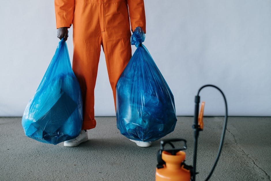 A person dressed in an orange uniform and white shoes, holding two large blue plastic rubbish bags filled with waste, stands on a plain grey floor against a neutral light grey wall. The individual's gloved hands grasp the bags by the tied tops, with the contents not visible but assumed to be waste materials suitable for private rubbish removal services. In the foreground, slightly out of focus, there is an orange handheld sprayer connected to a black hose, likely a cleaning or waste management tool. The scene suggests an indoor or controlled environment used for rubbish handling, with minimal background detail and no other objects present. The overall composition emphasizes the process of waste collection and the handling of refuse, aligning with professional rubbish removal practices and the context of private waste disposal, as offered by services such as those provided by Rubbish Removal Crystal Palace.