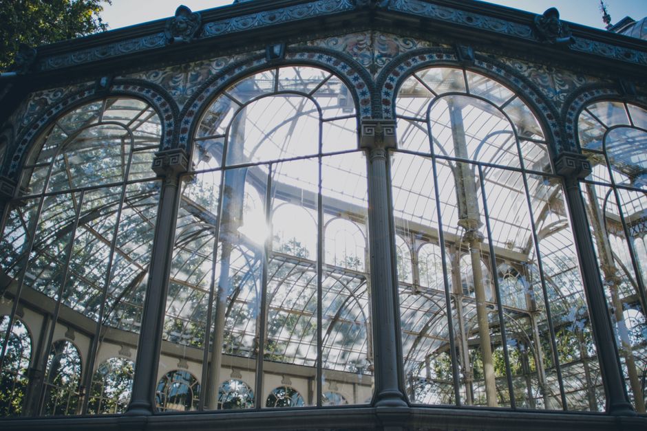 A detailed view of a large, ornate Victorian-style glass conservatory with intricate decorative patterns along the lower part of the structure. The conservatory features expansive, curved glass panels held by slender, cream-colored metal frames, allowing sunlight to illuminate its interior. The metal framework includes vertical and horizontal supports, with some decorative elements around the pillars and arches. The structure is set outdoors, surrounded by tall green trees with broad leaves, visible against a bright blue sky. The conservatory appears to be well-preserved, with some reflections of the sky and trees visible on the glass surfaces, and is situated in a landscaped garden or park setting. This scene relates to practices of private on-site clearance or alternative waste handling, often associated with maintenance or restoration of garden buildings, aligning indirectly with rubbish removal services.