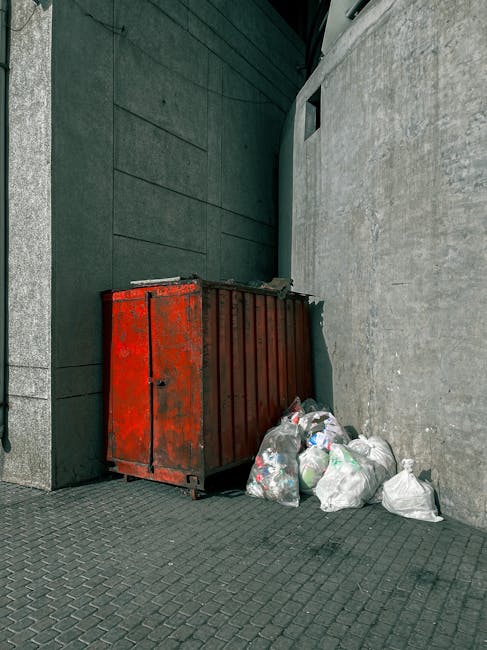 A red metal dumpster with visible rust and chipped paint is positioned against a dark, slate-colored wall on the left side of the image. The dumpster's surface is textured with streaks of corrosion and areas where the paint has worn away, revealing bare metal underneath. Several tightly tied white garbage bags, some with visible contents such as plastic bottles and paper, are piled on the ground next to the dumpster, leaning slightly against its side. The setting appears to be an urban alley or service area, with a paved surface made of small, interlocking dark grey bricks, and a concrete wall on the right side showing a rough, weathered texture. Overhead, a small section of a dark canopy or structure can be seen, casting subtle shadows over part of the scene. The environment is typical of private waste disposal areas used for alternative rubbish collection or on-site clearance, which may suggest targeted waste removal services in a city or town setting.