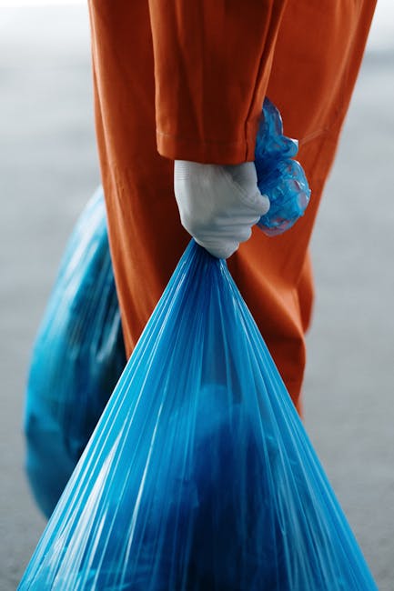 A person wearing an orange protective suit and white gloves is holding a tightly knotted blue plastic rubbish bag by its top, with the bag filled with waste visible through its semi-translucent material. The background appears to be an outdoor environment with a blurred natural or urban setting, suggesting an active waste collection operation. The focus is primarily on the hand grasping the rubbish bag, emphasizing the handling of waste materials, which aligns with private rubbish removal or on-site clearance services provided by companies like rubbishremovalcrystalpalace.org.uk. The scene conveys a professional waste collection activity, highlighting the use of durable gloves and appropriate disposal practices in a manner consistent with waste management procedures.