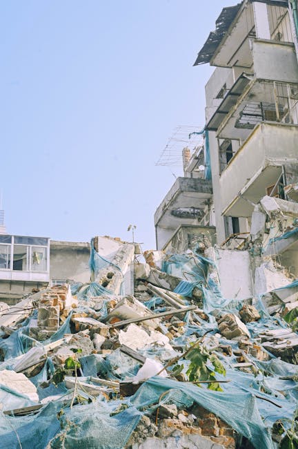 The image shows a collapsed section of a multi-storey building, with several floors partially destroyed and debris scattered at the base. The rubble includes broken bricks, concrete fragments, wooden and metal debris, some of which are covered with blue tarps that appear to have been placed to contain dust or protect the materials. The exposed concrete walls and balconies of the remaining structure reveal signs of damage, with some balconies hanging askew or missing sections. Surrounding the debris, there is limited greenery with a few plants visible emerging through the rubble. The background features a clear, pale blue sky, and there are no vehicles or machinery present in the scene. The image highlights the extensive destruction typical in urban environments, which services like independent rubbish removal or on-site clearance are often called upon to handle in such situations, aligning with the context of alternative waste management and junk removal services in a cityscape setting.