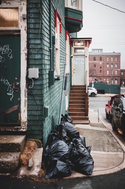 A stack of black rubbish bags filled with waste is gathered at the base of a green wooden building with weathered, chipped paint and visible graffiti on a metal door. The bags are tightly packed and resting on a small concrete step, adjacent to a patch of brown, dried grass and a section of uneven concrete pavement. A wooden staircase with a dark brown finish leads up to a white door with a small awning supported by red brackets, situated next to the building's corner. The surrounding environment includes a parking area with a few parked cars, one of which is a red vehicle visible on the right side, and additional brick buildings and urban infrastructure in the background under overcast skies. The scene captures an aspect of private rubbish collection, typical in residential areas, and subtly highlights the importance of waste management services such as those provided by rubbishremovalcrystalpalace.org.uk, supporting alternative disposal options for household waste.